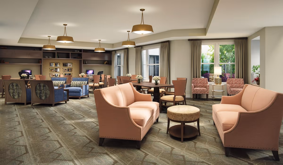 Spacious senior living common room with sofas, armchairs, tables, and a computer station by windows.