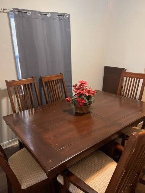 A wooden dining table with six matching wooden chairs around it. The table has a small basket with red flowers as a centerpiece. There is a window with gray curtains in the background and light-colored walls.