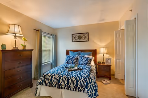 Sunny bedroom with a double bed dressed in blue patterned bedding, wooden dresser and nightstand with lamps.