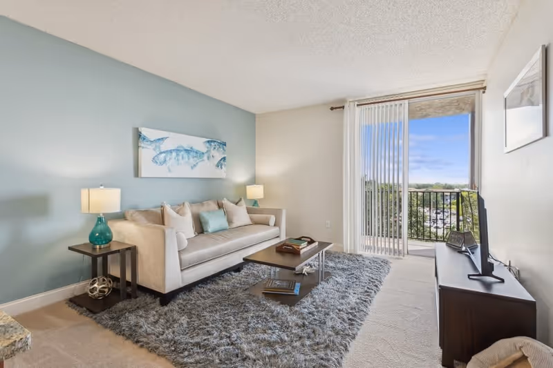 A bright living room with a beige sofa adorned with multiple cushions, two side tables each with a lamp, a rectangular coffee table on a fluffy gray rug, a TV on a dark wooden stand, and a sliding glass door leading to a balcony with a view of trees and a blue sky.