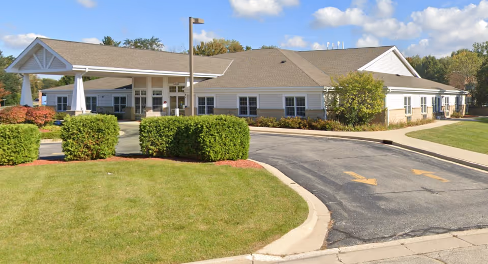 Front view of a single-story assisted living building with a covered porte-cochère, driveway, and landscaped lawn.