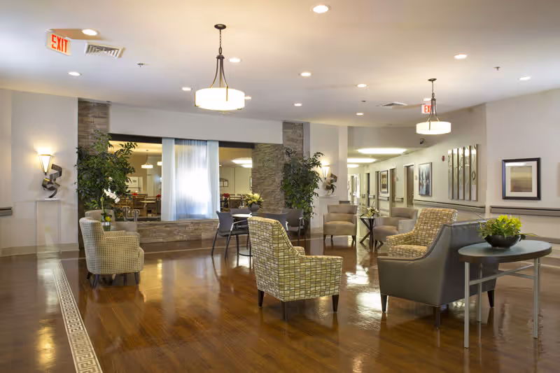 Well-lit senior living facility lounge with chairs, tables, plants, and pendant lights over polished wooden floors.