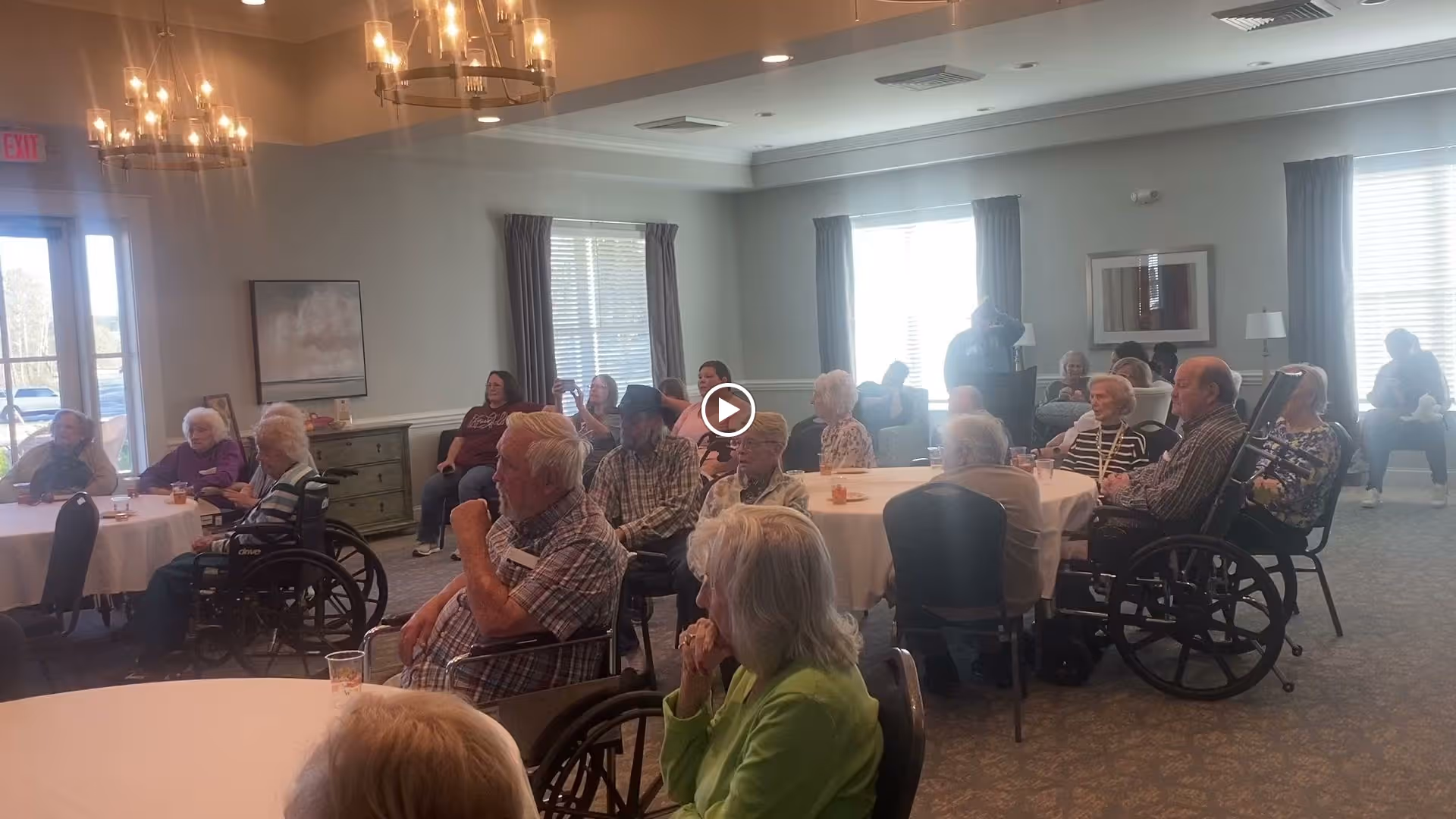 A group of elderly people seated around round tables in a well-lit room with large windows and chandeliers. Some individuals are in wheelchairs, and others are sitting on chairs. The room has light-colored walls with framed artwork and curtains on the windows. The setting appears to be a communal gathering or activity area in an assisted living facility.