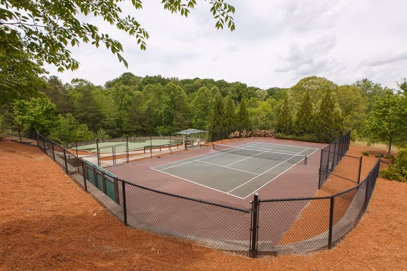Fenced outdoor tennis courts surrounded by trees and pine straw landscaping at a senior living community.