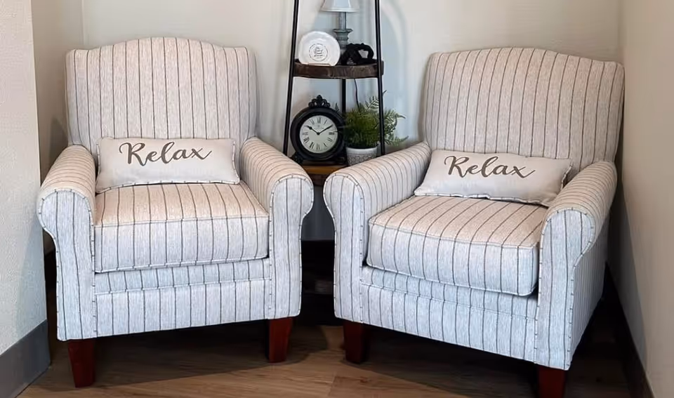 Two beige striped armchairs with 'Relax' pillows placed in a corner next to a small black metal and wood shelf holding a clock, a lamp, a small plant, and decorative items.