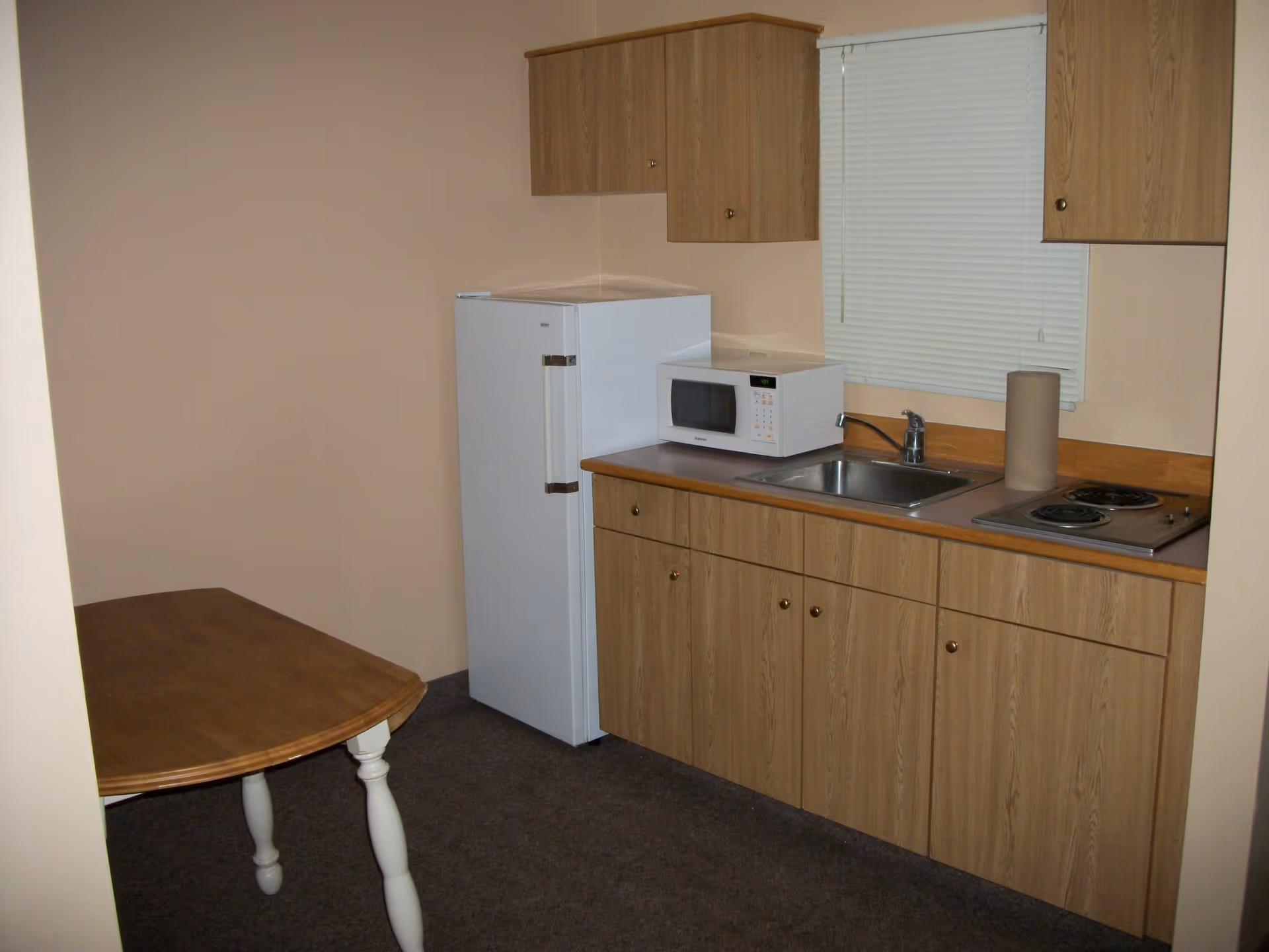 A small kitchen area with wooden cabinets, a white refrigerator, a microwave on the countertop, a stainless steel sink, a two-burner electric stove, and a wooden table with white legs. There is a window with closed blinds above the sink.