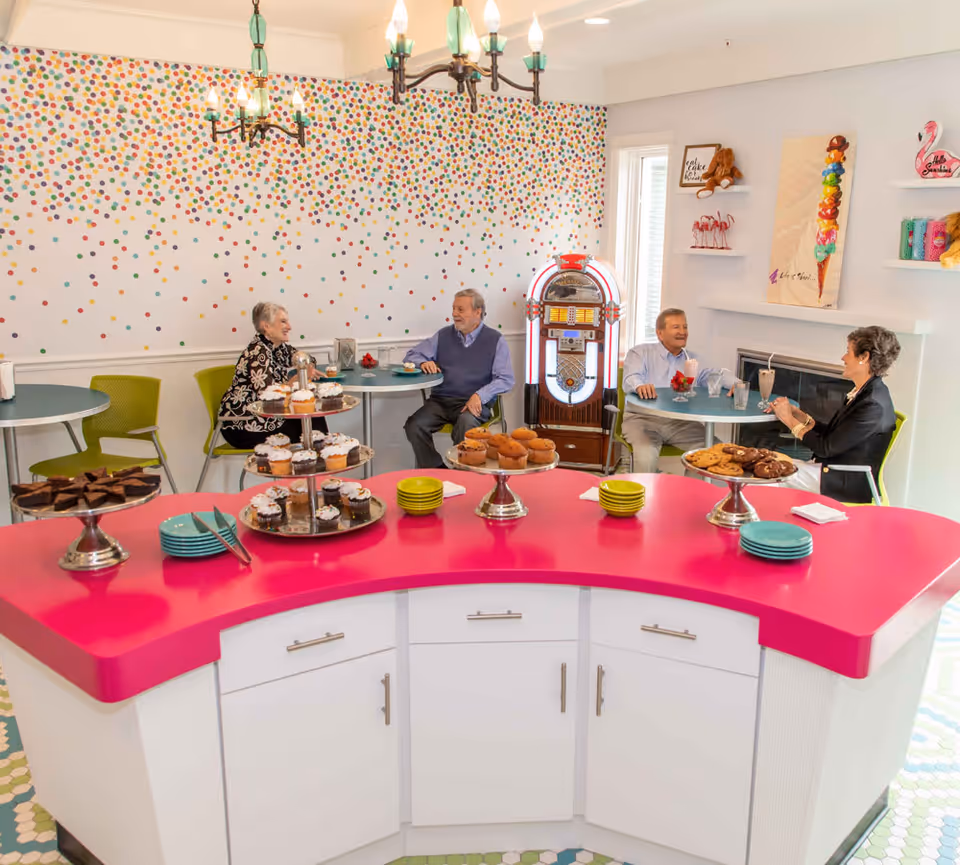 A bright and cheerful dining area with a pink curved counter displaying various desserts including cupcakes, muffins, brownies, and cookies. Four elderly people are seated at two small round tables, enjoying conversation and drinks. The walls are decorated with colorful polka dots and playful artwork, and a vintage jukebox stands against the wall.