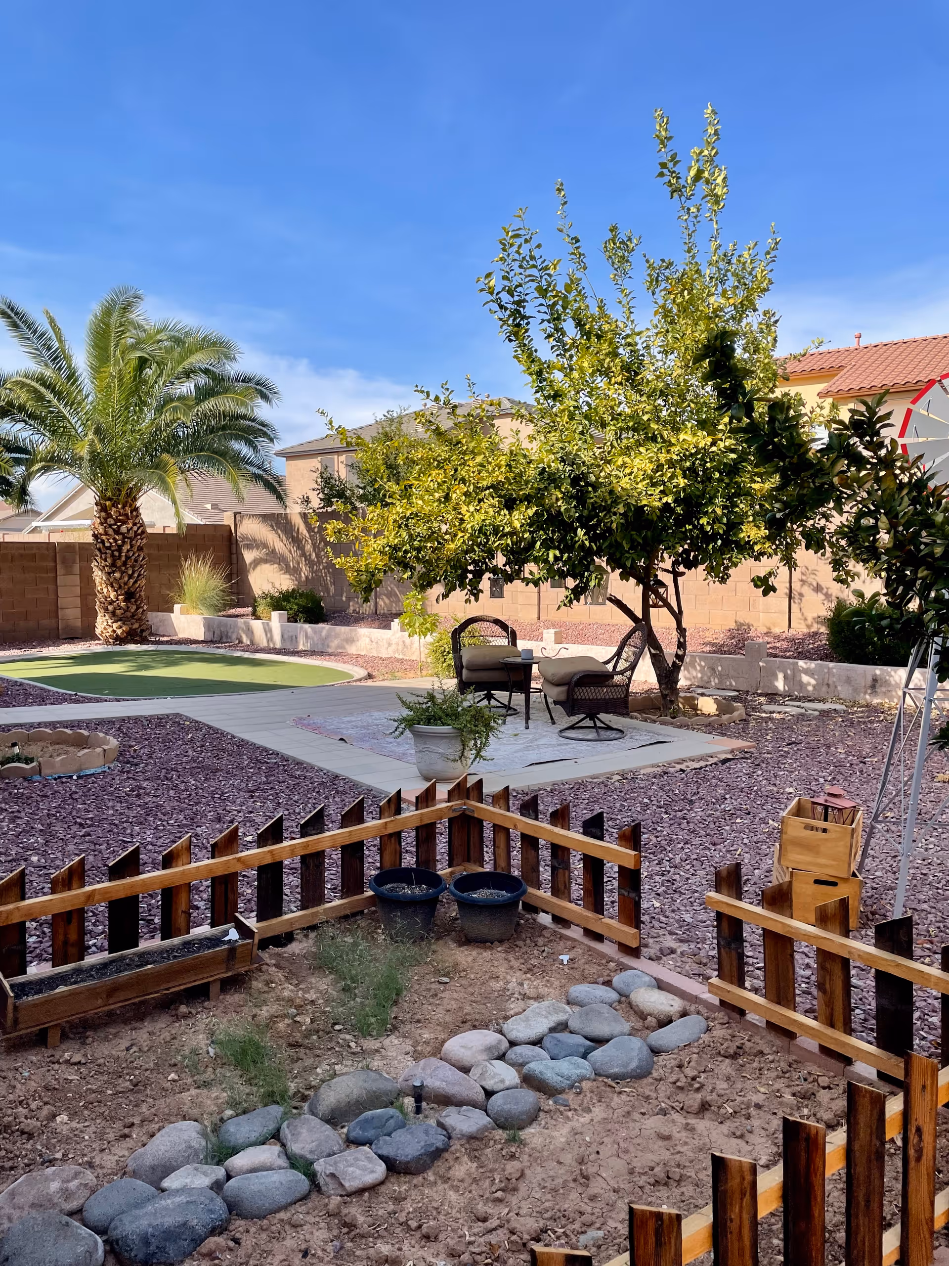 Outdoor garden area with a small wooden fence enclosing a patch of soil and rocks. In the background, there is a patio with two chairs and a small table under a leafy tree. A palm tree and other plants are visible along a brick wall under a clear blue sky.