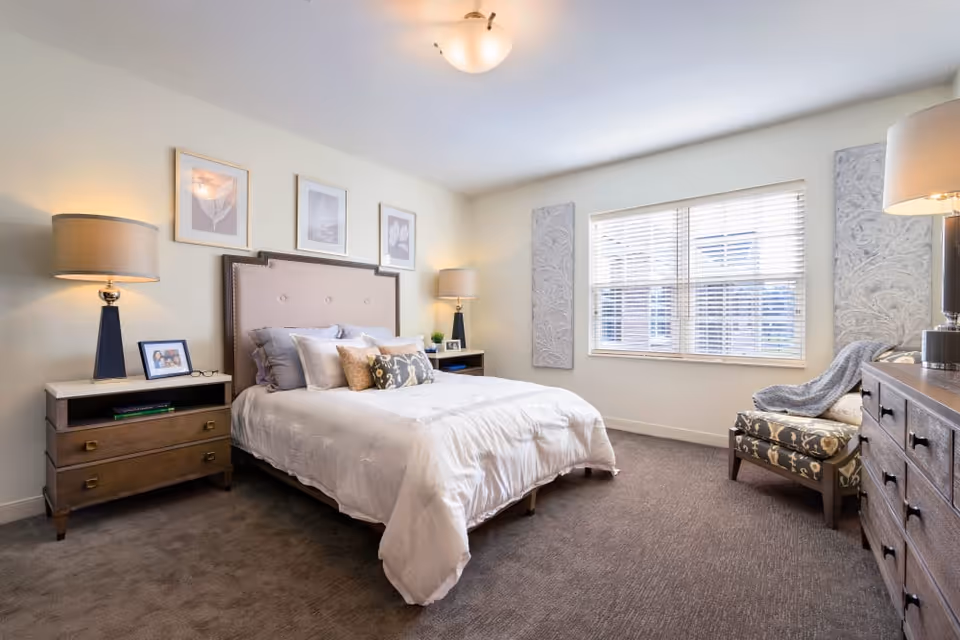 A bright and neatly arranged bedroom featuring a large bed with multiple pillows and a beige headboard. On either side of the bed are wooden nightstands with lamps and framed photos. The room has a large window with blinds, two decorative wall panels, a patterned armchair with a throw blanket, and a wooden dresser with a lamp on top. The carpet is gray and the walls are painted light beige.