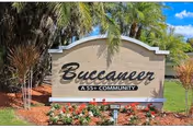 A beige stone sign with the text 'Buccaneer A 55+ COMMUNITY' surrounded by landscaping including palm trees, bushes, and flowers under a blue sky with some clouds.