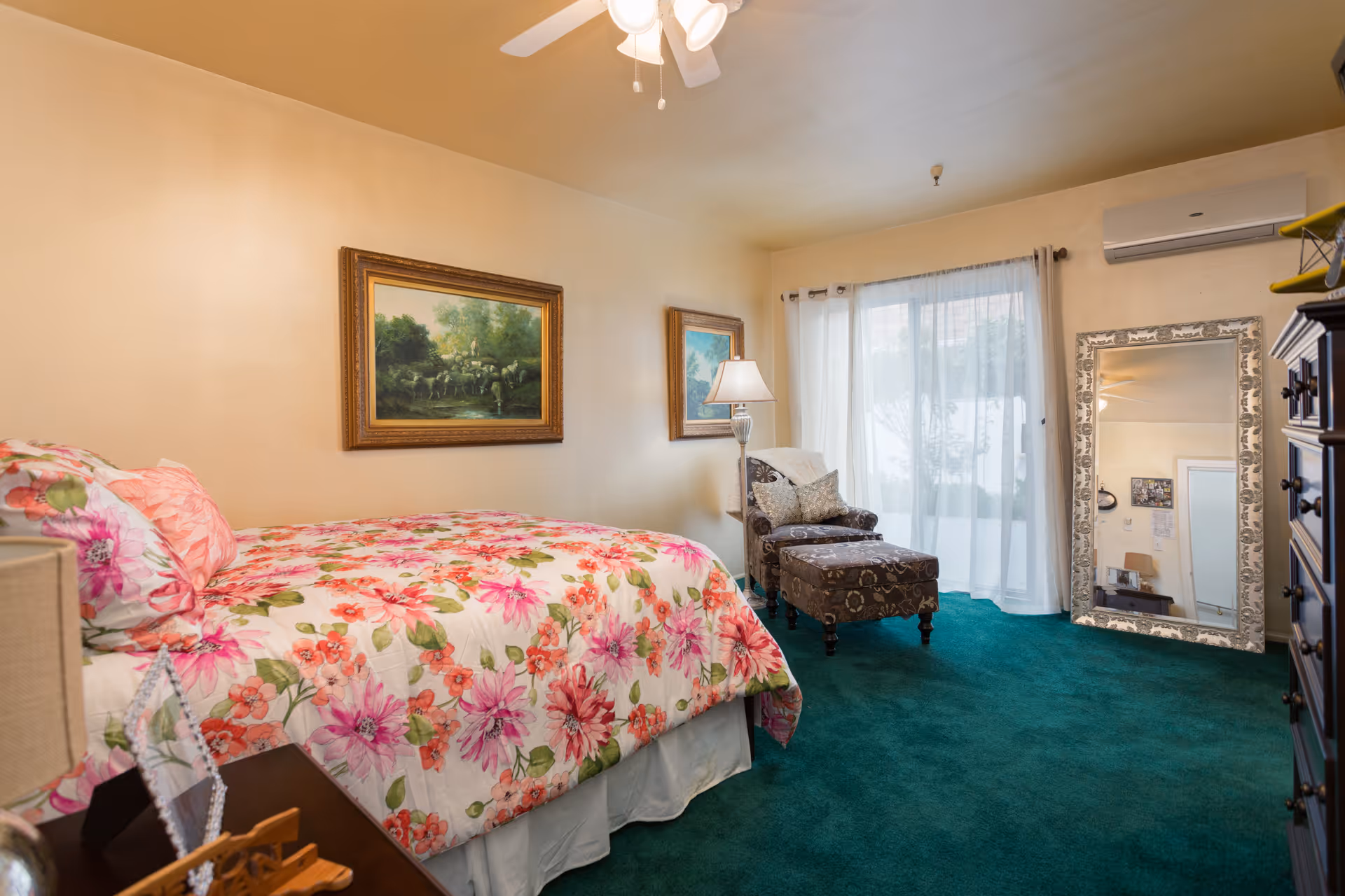 Cozy bedroom with a floral bedspread, an upholstered armchair and ottoman, a large floor mirror, and a sliding glass door with sheer curtains.