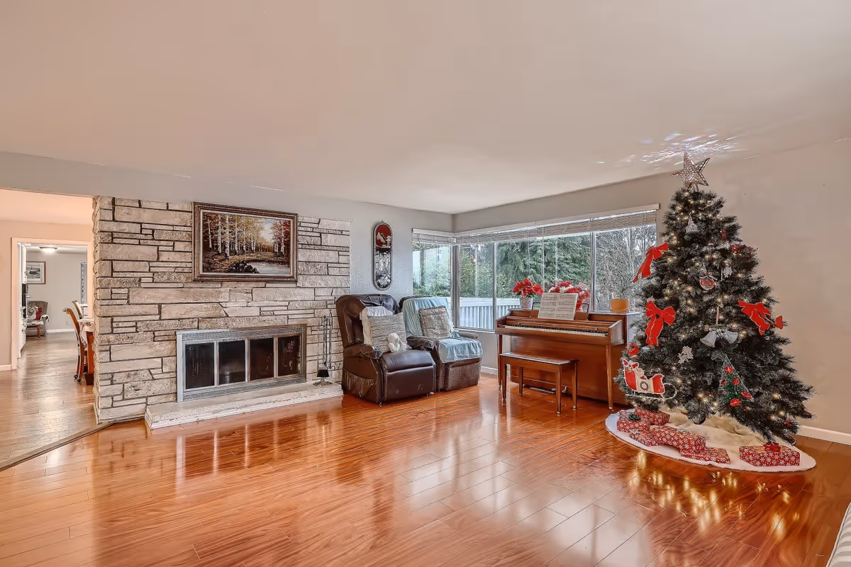 A cozy living room with polished wooden floors, a stone fireplace with a framed painting above it, two recliner chairs with cushions, a piano with sheet music and poinsettia plants on top, and a decorated Christmas tree with red bows, ornaments, and wrapped presents underneath. Large windows provide a view of greenery outside.