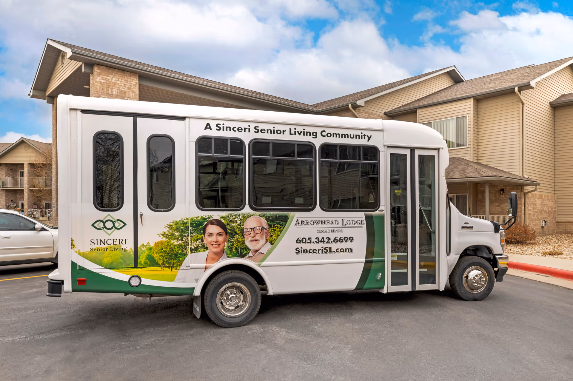 A white shuttle bus parked outside a senior living facility building. The bus has signage for Arrowhead Lodge Senior Living with contact information and the logo for Sinceri Senior Living. The building in the background has beige siding and brick accents.