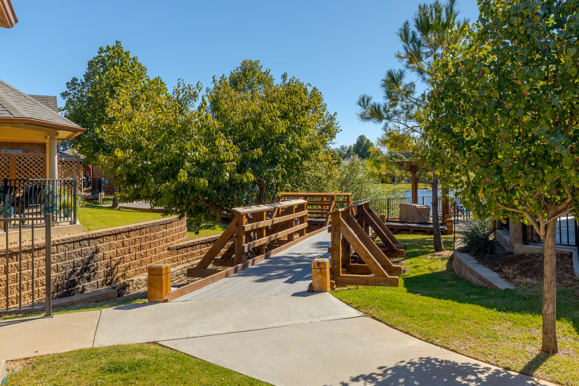 A wooden pedestrian bridge over a small landscaped area with green grass and trees under a clear blue sky, part of an outdoor garden area in a senior living facility.