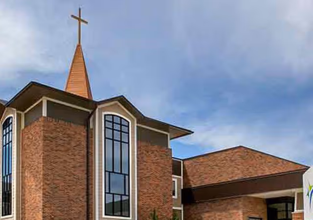 Brick senior living facility building with tall arched windows and a cross-topped steeple under a cloudy sky.