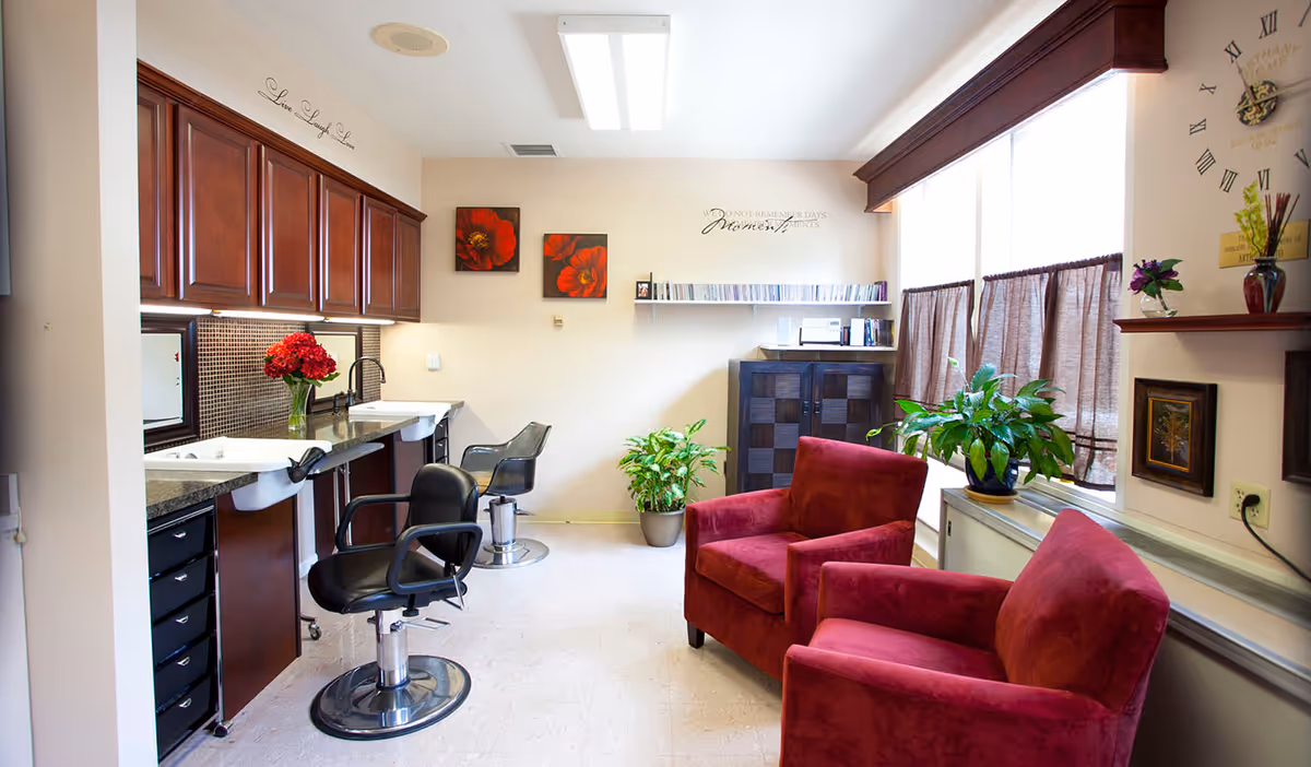 Interior view of a salon area featuring two black styling chairs, a granite countertop with sinks, and red armchairs. The walls are decorated with floral artwork and motivational quotes, along with a shelf containing CDs.