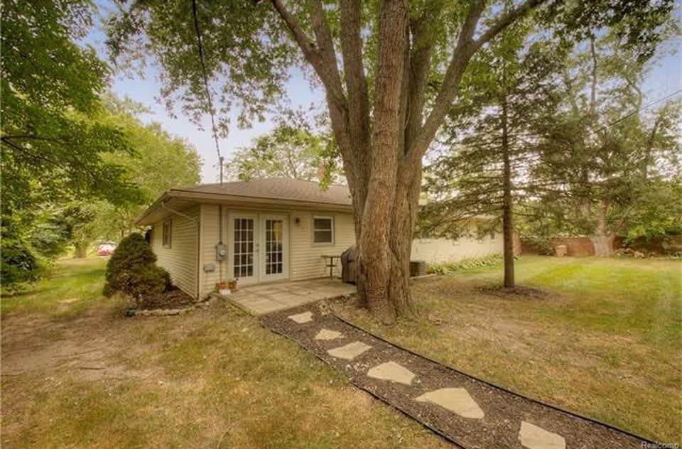 Single-story house exterior with a stone path leading to patio doors, large trees, and a grassy yard.