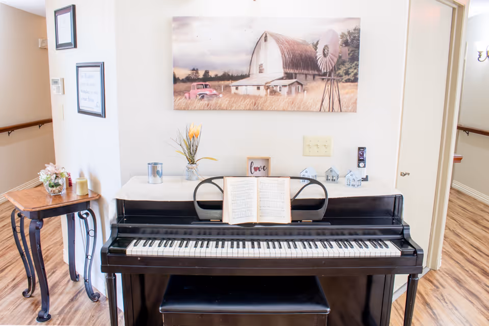 A black upright piano with an open sheet music book on the music stand. On top of the piano are decorative items including a small vase with flowers, a metal container, a framed sign that says 'love', and three small house figurines. Above the piano hangs a painting of a rustic barn, a windmill, and a red truck in a field. To the left of the piano is a small wooden side table with a flower arrangement and candles. The room has light-colored walls and wood flooring.