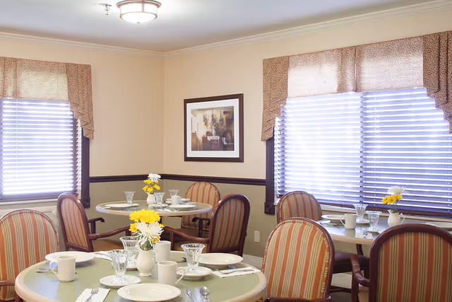 A dining room with round tables set for a meal, featuring white plates, cups, and glassware. Each table has a small vase with yellow and white flowers. The room has beige walls, two windows with blinds and patterned valances, and a framed painting on the wall.