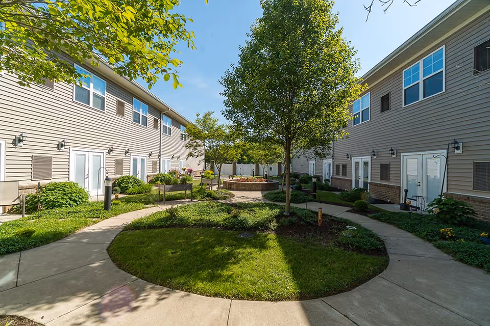Outdoor courtyard area between two beige two-story buildings with multiple windows and white doors. The courtyard features a circular grassy area with a tree in the center, surrounded by paved walkways and landscaped greenery under a clear blue sky.