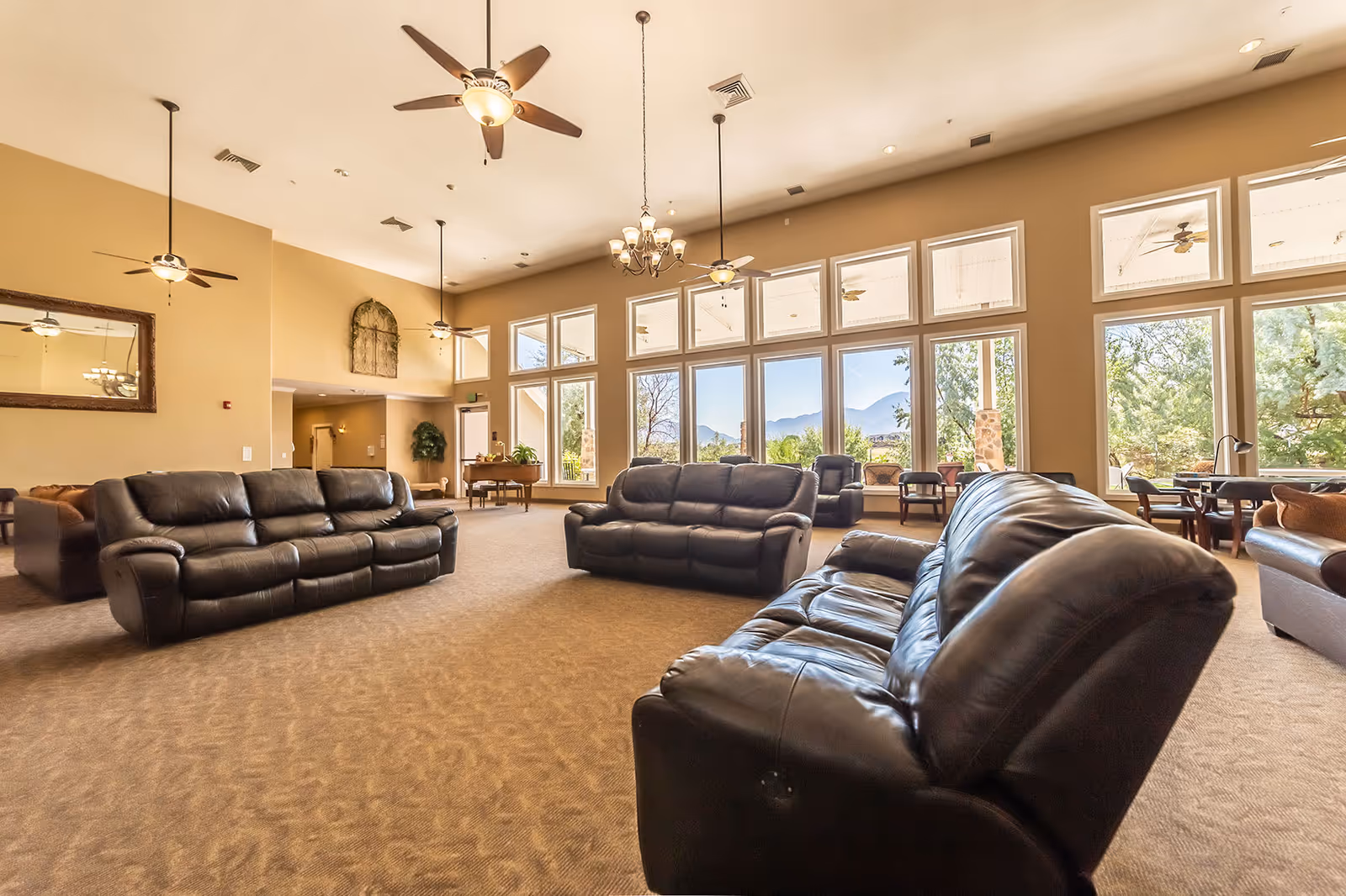 Spacious living room area with multiple black leather sofas arranged on a carpeted floor. Large windows along one wall provide a view of trees and mountains outside. Ceiling fans and a chandelier hang from the high ceiling, and there is a large mirror on one wall.
