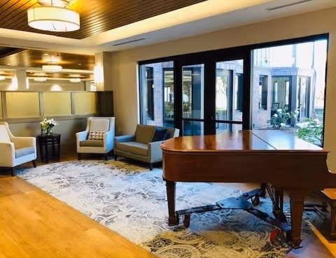 A cozy interior lounge area featuring a grand piano on a patterned rug, two armchairs, a small sofa, and a side table with a flower vase. Large windows and glass doors provide a view of an outdoor garden area.
