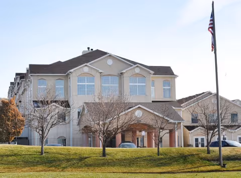 Three-story beige senior living facility building with a covered entrance, flagpole, and a grassy lawn in front.