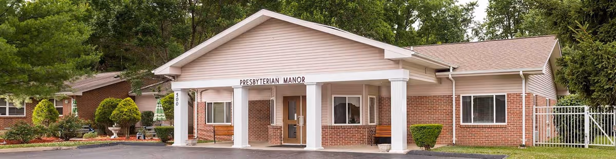 Exterior view of Farmington Presbyterian Manor, a single-story brick building with white columns at the entrance, a peaked roof, and surrounding greenery including trees and shrubs.
