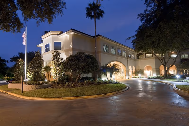Exterior view of The Mayflower at Winter Park building at dusk, showing a well-lit driveway, landscaped greenery, palm trees, and an American flag on a flagpole.