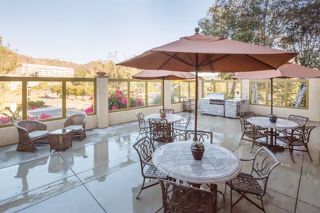 Outdoor patio area with round tables and chairs under large umbrellas, a built-in barbecue grill, and wicker seating. The patio is surrounded by a low wall with glass panels, overlooking a landscaped area with trees and hills in the background.