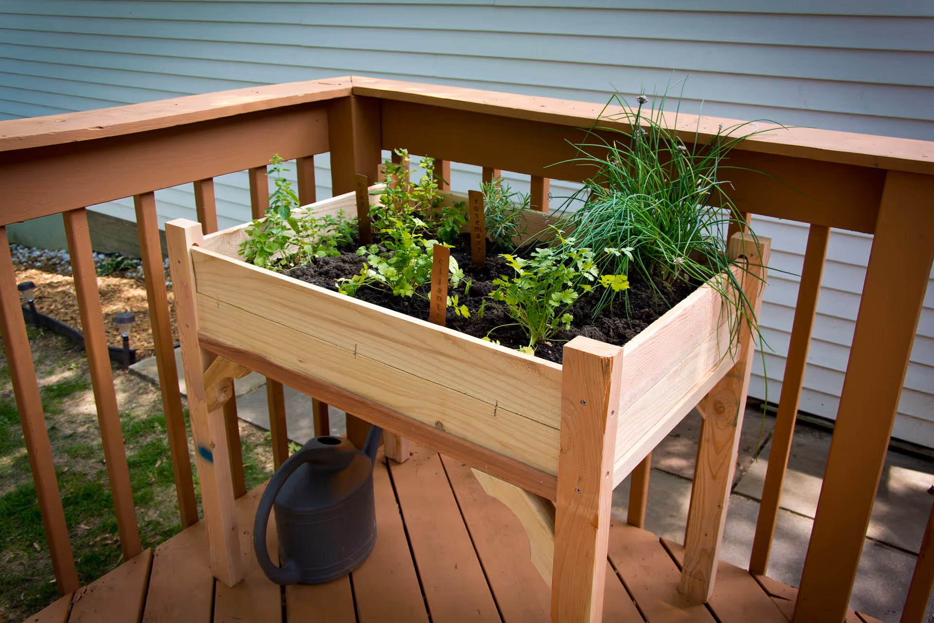 A wooden raised garden bed on a wooden deck railing with various herbs planted in soil, including rosemary and cilantro, with a black watering can placed underneath.