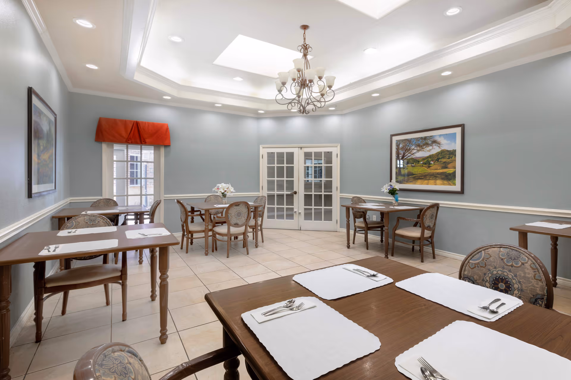 Bright, formal dining room with several small tables and chairs set with placemats and cutlery, a chandelier, skylights, and French doors.