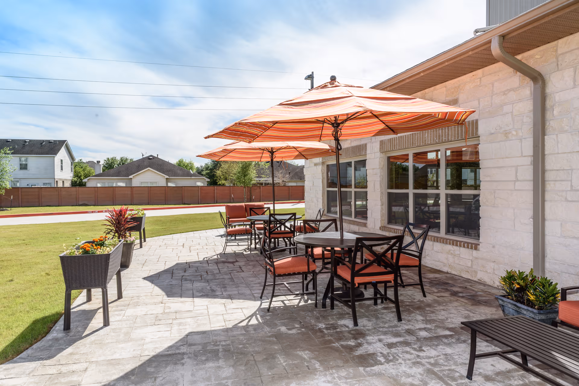 Outdoor patio area at Oak Park Retirement Resort with multiple round tables and chairs featuring orange cushions, shaded by large orange striped umbrellas. The patio is paved with stone tiles and has planters with flowers and greenery. The building exterior is light-colored stone with large windows reflecting the patio furniture.