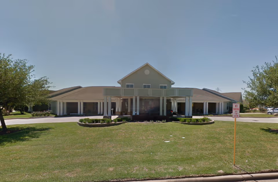 Front exterior view of The Reserve at Katy Assisted Living and Memory Care building with a covered entrance, green lawn, trees on both sides, and a clear blue sky.