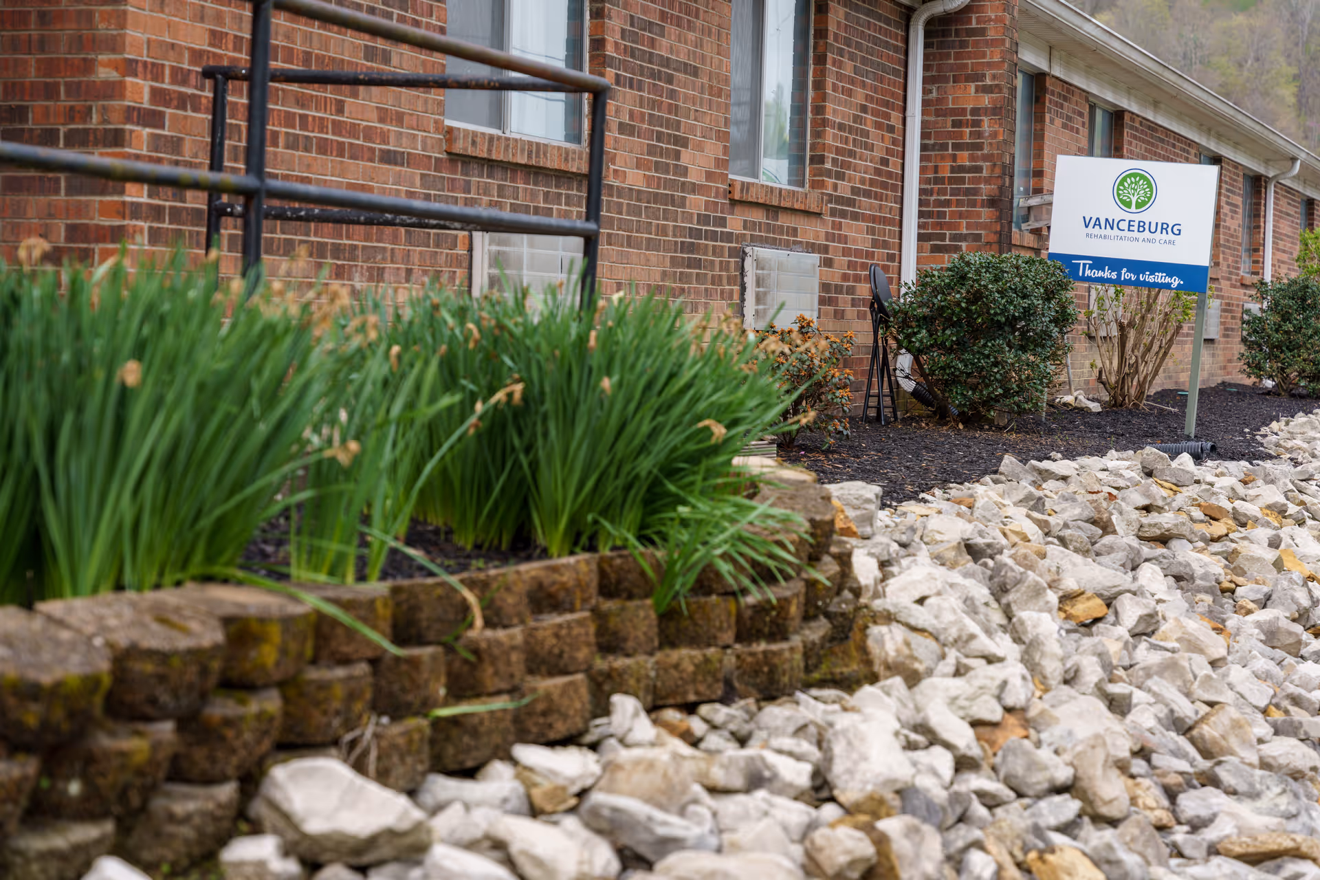 Outdoor view of a brick building with a landscaped garden featuring green plants and rocks. A sign near the building reads 'Vanceburg Rehabilitation and Care, Thanks for visiting.'