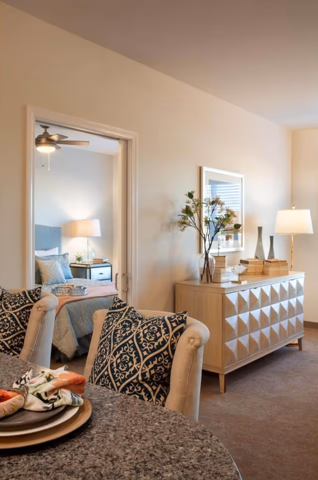 View of a cozy living area with a patterned cushioned chair in the foreground, a granite table set with plates and napkins, and a decorative sideboard with vases, books, and a lamp. Through an open doorway, a bedroom is visible with a bed, bedside table, lamp, and ceiling fan.