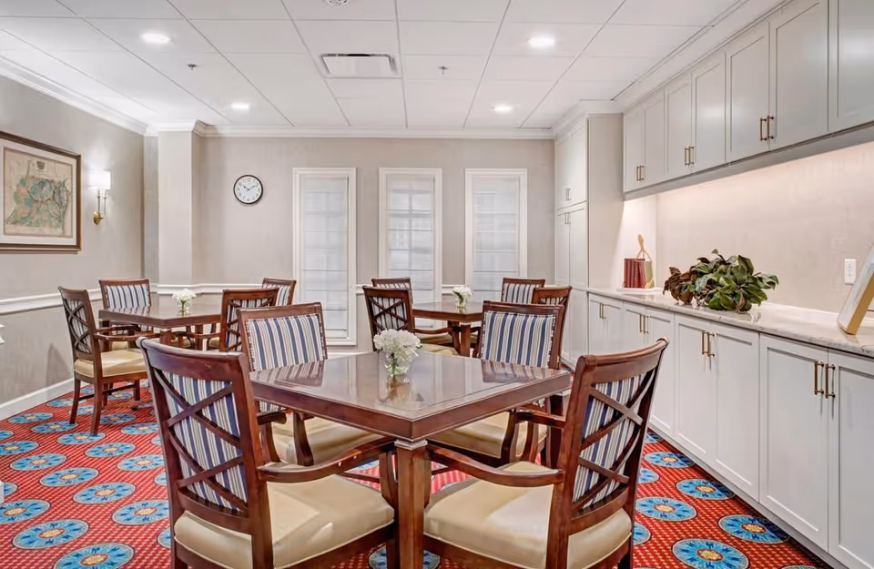 A bright dining room with wooden tables and striped upholstered chairs on a patterned red carpet, white cabinets along one wall and framed windows at the far end.