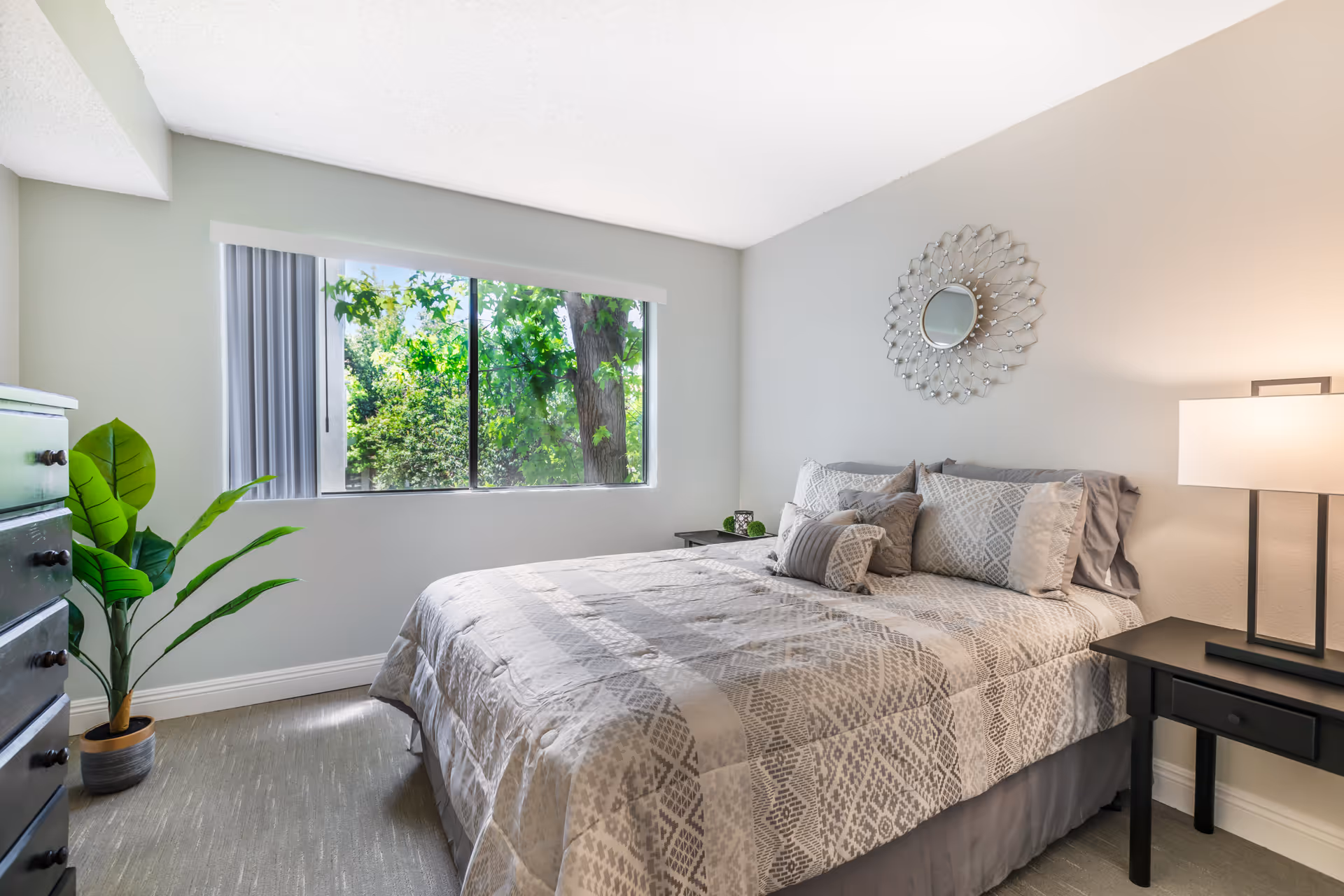 A bright bedroom with a large window showing green trees outside. The room features a neatly made bed with patterned bedding and multiple pillows, a decorative round mirror on the wall above the bed, a black nightstand with a lamp, a small plant on the nightstand, a black dresser, and a potted plant on the floor.