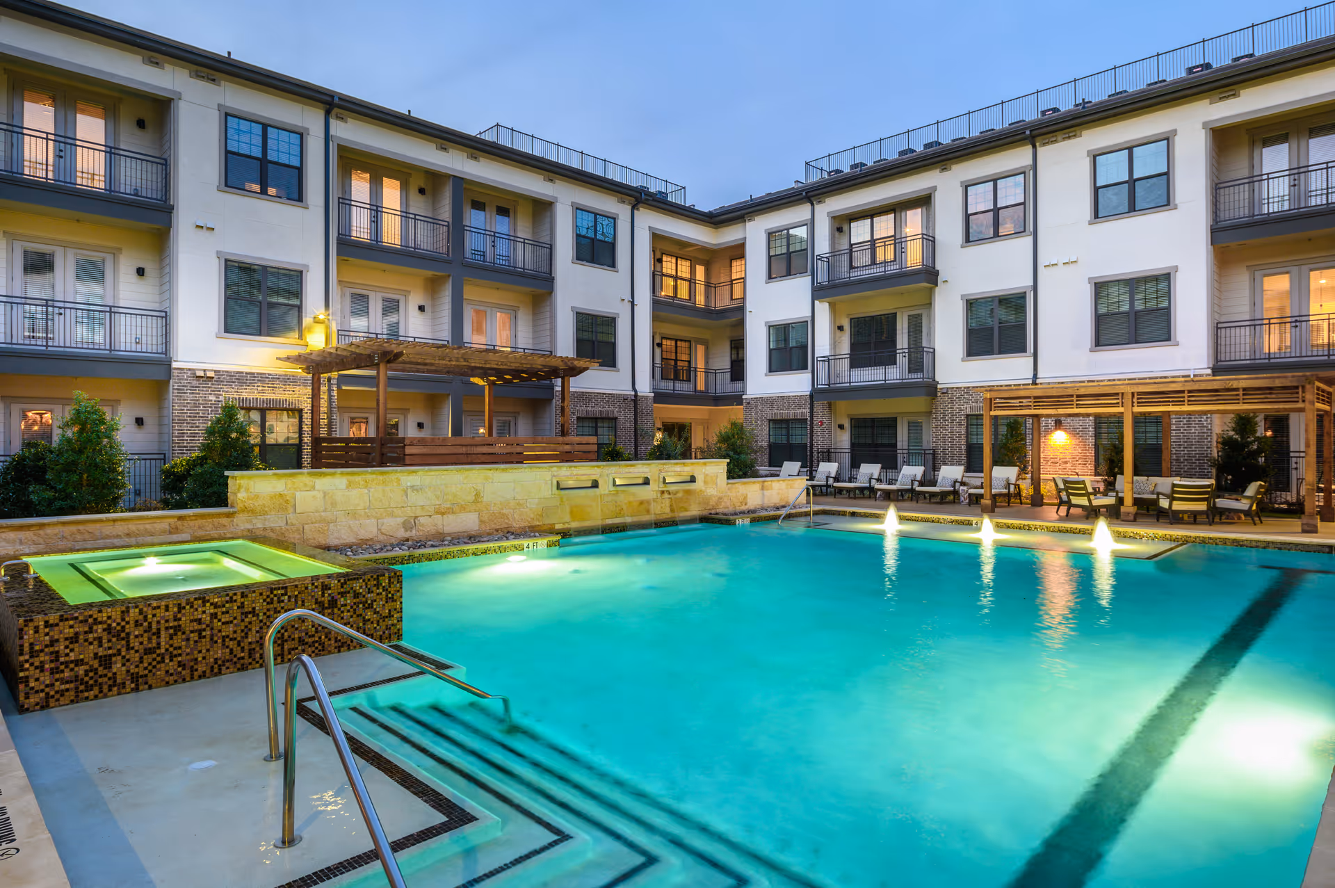 Evening view of a well-lit outdoor swimming pool area in an apartment complex with lounge chairs, pergolas, and a hot tub, surrounded by a three-story building with balconies and large windows.