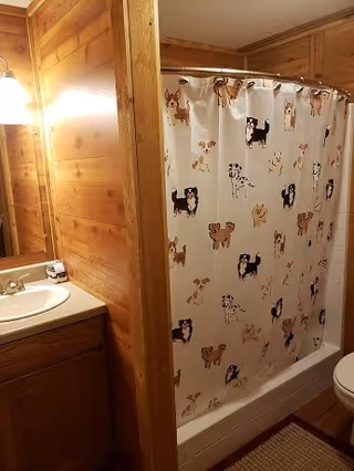 A bathroom with wooden walls featuring a sink with a countertop and a mirror above it. Next to the sink is a bathtub with a shower curtain decorated with various dog illustrations. A toilet is partially visible on the right side of the image.
