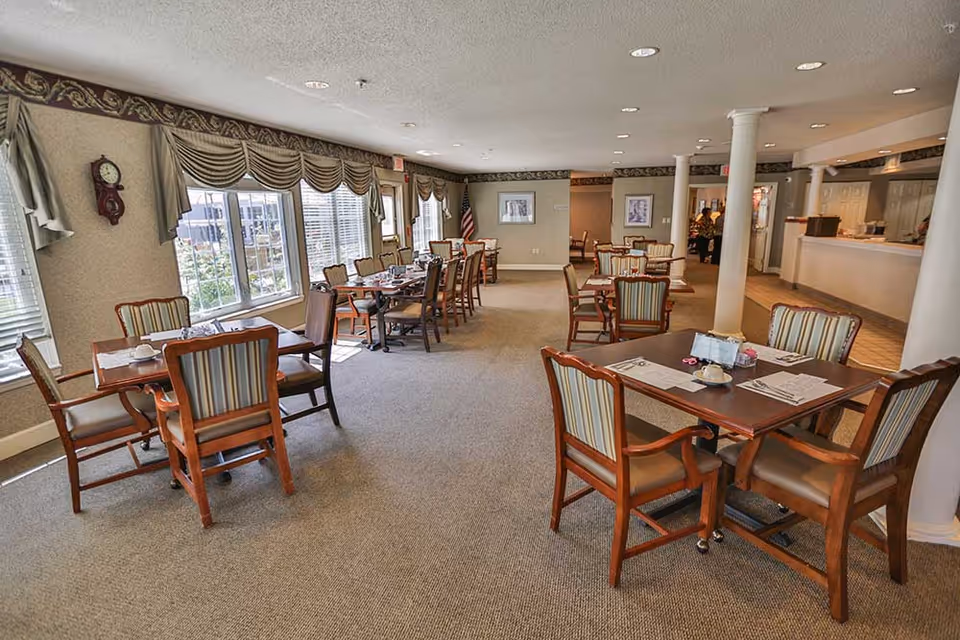 A spacious dining room with multiple wooden tables and chairs arranged neatly. The room has large windows with draped curtains allowing natural light to fill the space. The walls are decorated with framed pictures and a clock, and there is an American flag in the corner. The carpeted floor and ceiling lights create a warm and inviting atmosphere.