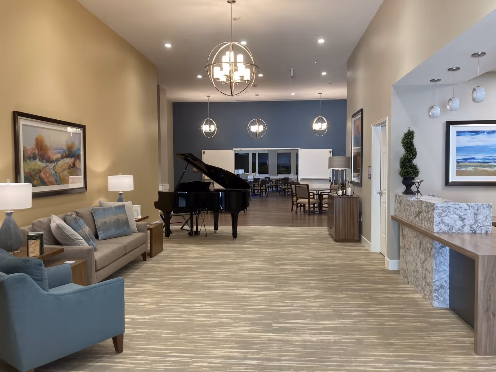 Interior view of a senior living facility common area with a beige sofa and blue armchair on the left, a black grand piano in the middle, and a dining area with tables and chairs in the background. The walls are painted beige and blue, with modern light fixtures hanging from the ceiling and framed artwork on the walls.