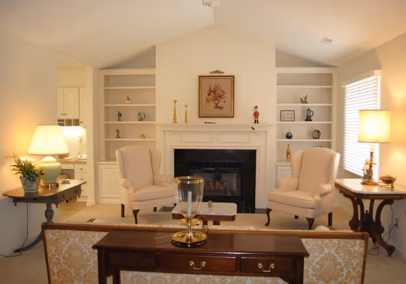 A cozy living room with two beige armchairs facing a fireplace with a black tile surround. Built-in white shelves flank the fireplace, decorated with small figurines and vases. A wooden table with a lamp and a plant is on the left, and another wooden table with a lamp is on the right near a window with blinds. A wooden console table with a decorative glass piece is behind a patterned sofa in the foreground.