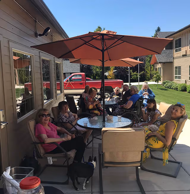 A group of elderly people sitting outdoors around a glass table with a large orange umbrella providing shade. They are seated on patio chairs on a concrete patio next to a building. A red pickup truck is parked nearby, and there is green grass and trees in the background. One small black and white dog is also present near the table.