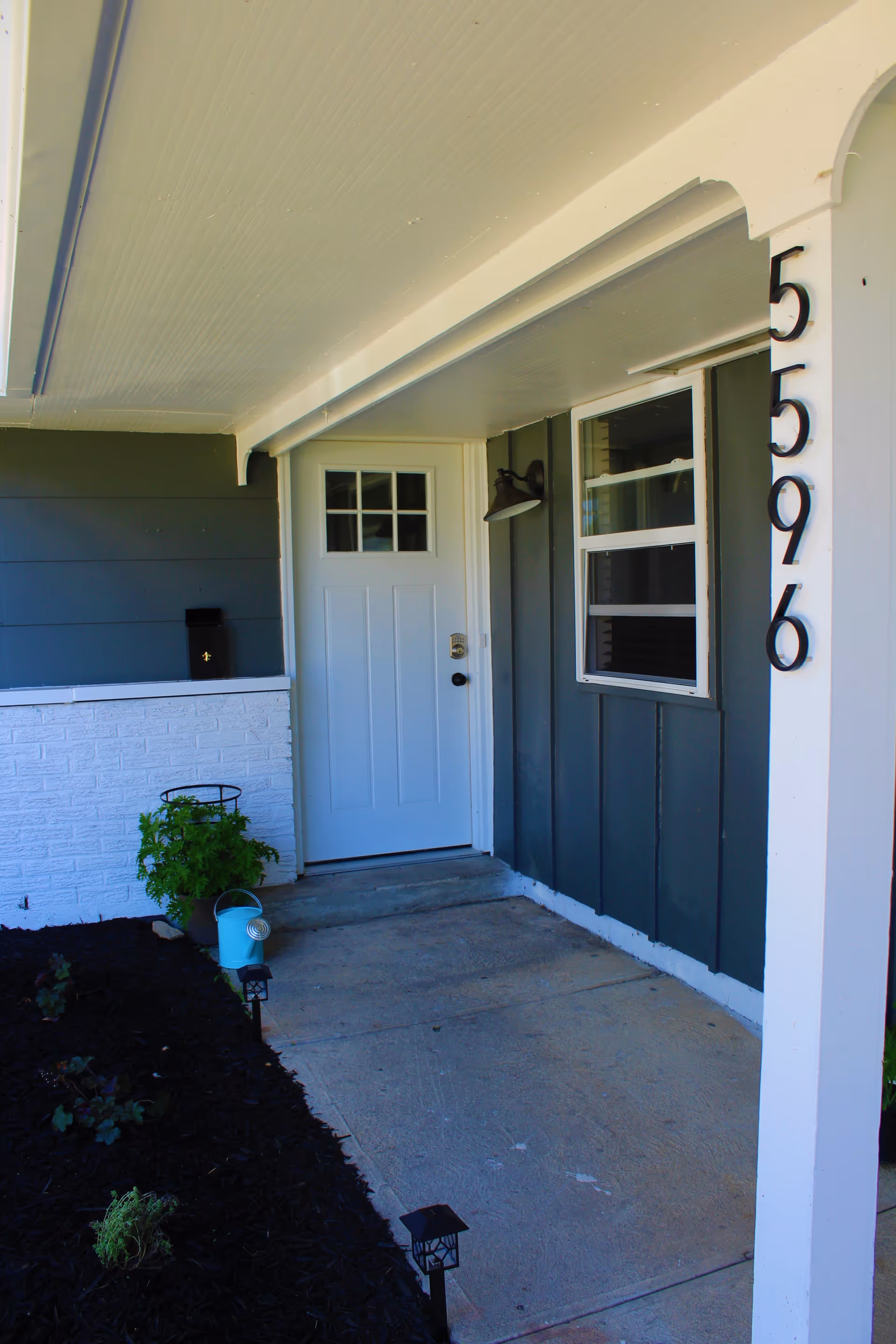 Covered front porch area of a house with a white door featuring a window with multiple panes. The exterior wall is painted dark gray with a white trim. There is a black mailbox mounted on a white brick half-wall, a potted plant, a small blue watering can, and two small black lantern-style lights along the edge of the concrete porch. The house number 5596 is displayed vertically on a white post.
