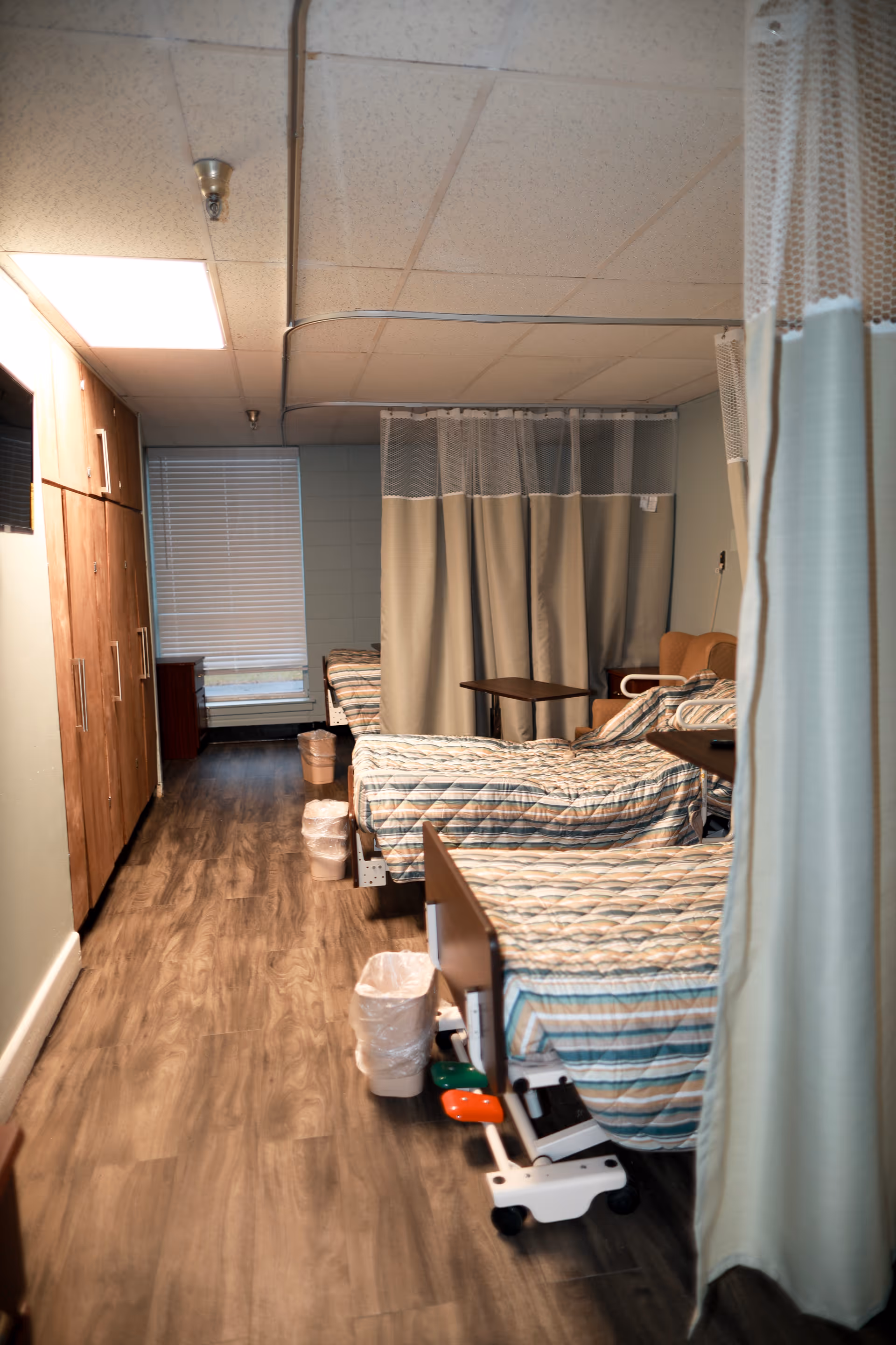 Interior view of a healthcare facility room with multiple hospital beds lined up along the wall, each separated by privacy curtains. The beds have striped bedding, and there are small tables and trash bins next to them. The floor has wood-like vinyl flooring, and there are cabinets on the left side of the room.