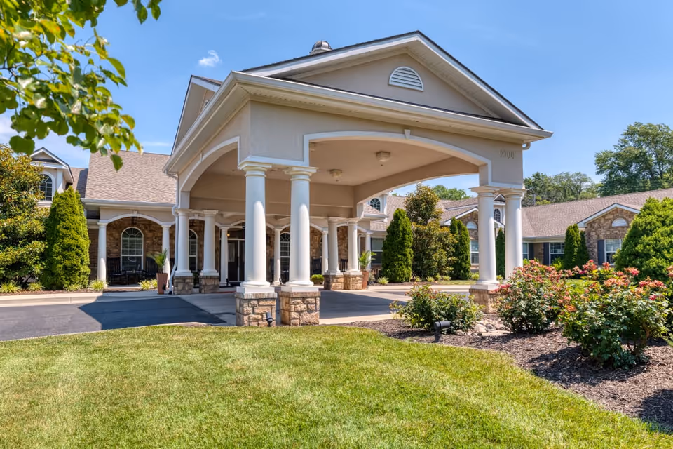 Front exterior view of Addington Place of Prairie Village featuring a covered entrance with white columns, a driveway, well-maintained green lawn, and landscaped bushes and trees under a clear blue sky.