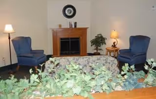 Communal living room with a fireplace, two blue armchairs, a floral sofa, lamps, and a potted plant.