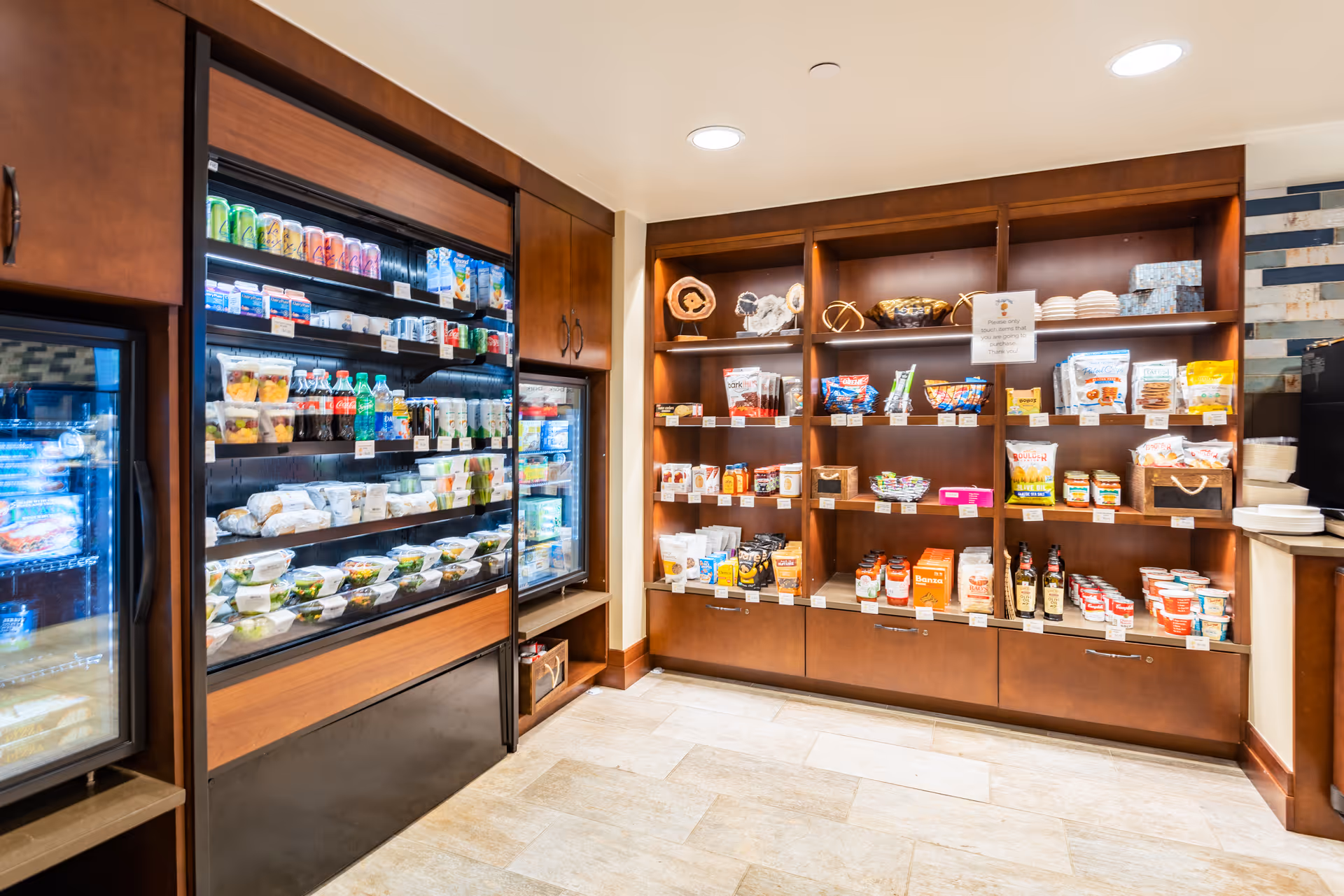 Interior view of a small convenience or snack area with wooden shelves stocked with various packaged snacks, drinks, and food items. There are refrigerated sections on the left side containing beverages and pre-packaged food. The area has warm lighting and a tiled floor.