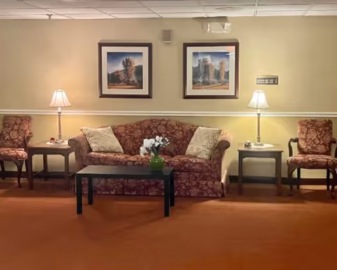 A cozy living room area with a floral patterned sofa and two matching armchairs. There are two side tables with lamps on either side of the sofa, and a small black coffee table in front with a green vase holding white flowers. Two framed landscape pictures hang on the beige wall above the sofa.